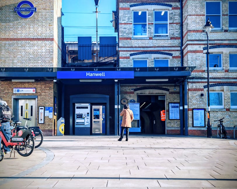 A person standing outside the Hanwell station entrance in Chadwell Heath, London, with a brick building façade featuring large windows and a blue signage board. The scene includes bicycles parked to the left and a covered ticket vending area with automated machines, as well as digital information displays on the walls. The station's entrance is located near a designated walkway on the pavement, which is being used in the context of home relocation or furniture transport, reflecting the logistical aspects of moving services offered by Man with Van Chadwell Heath.