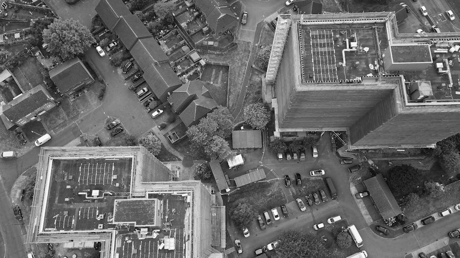 An aerial view showing a city scene with residential and commercial buildings, including a large multi-storey office building with solar panels on the roof. The image depicts a parking area with numerous parked cars, some lined up along the pavement and others within designated spaces. Adjacent to the buildings, there are small trees and a few shed structures, likely used for storage or utility purposes. Outside, on the pavement, a man wearing a high-visibility vest is seen lifting or carrying a cardboard box, part of a house removal or moving process. Nearby, a large moving van, branded as Man with Van Chadwell Heath, is parked close to the property, with the loading area adjacent to the entrance. The van is equipped for furniture transport and packing, with visible protective blankets and straps securing items. The setting suggests an organized home relocation involving packing, furniture transport, and loading activities, supported by the company's removal services in the Barking and Dagenham area.