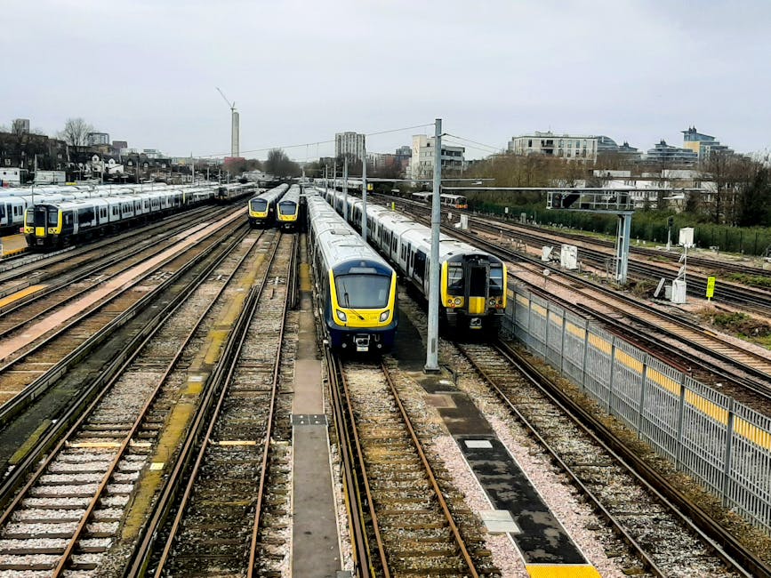 A view of multiple trains parked at a railway station with several parallel tracks, including a modern yellow and grey train in the foreground and other trains on adjacent tracks. Overhead electrical wires and supporting poles run above the trains. In the background, there are various buildings and residential apartments, with some trees and greenery visible. The scene is overcast, with grey skies casting diffused light over the station. The railway platform is not visible, but the trains are positioned close to the edge of the tracks, suggesting a loading and unloading area suited for home relocation and furniture transport services. This setting exemplifies the logistics involved in moving house contents via rail, supported by transportation companies like Man with Van Chadwell Heath, who coordinate loading, transit, and delivery processes in urban environments.