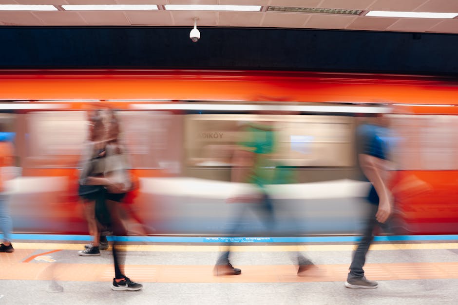 Image shows several passengers standing on a subway platform as a train with an orange and gray exterior rapidly passes by, creating a motion blur effect. The passengers are wearing casual clothing, some holding bags or backpacks, and appear momentarily stationary while waiting or preparing to board. The platform features safety markings in blue and yellow near the edge. The lighting indicates an indoor, underground station environment. This scene illustrates typical public transport conditions, relevant to home relocation and moving logistics when coordinating transport routes and timing for house removals, as handled by companies like Man with Van Chadwell Heath.