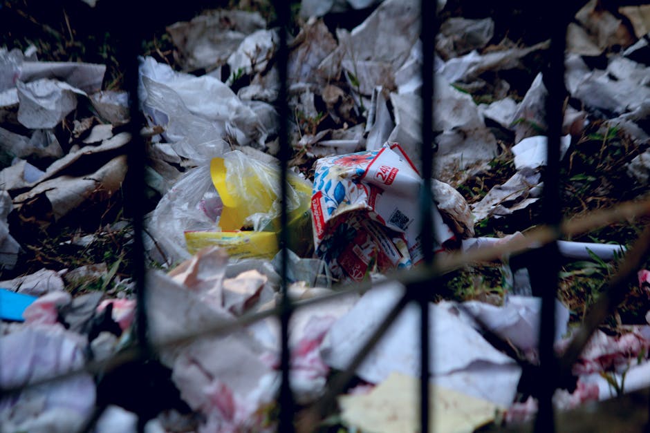 Close-up of a person wearing orange protective overalls and a white glove, holding a large, blue plastic bag filled with waste or recyclable materials. The background shows an outdoor setting with a p