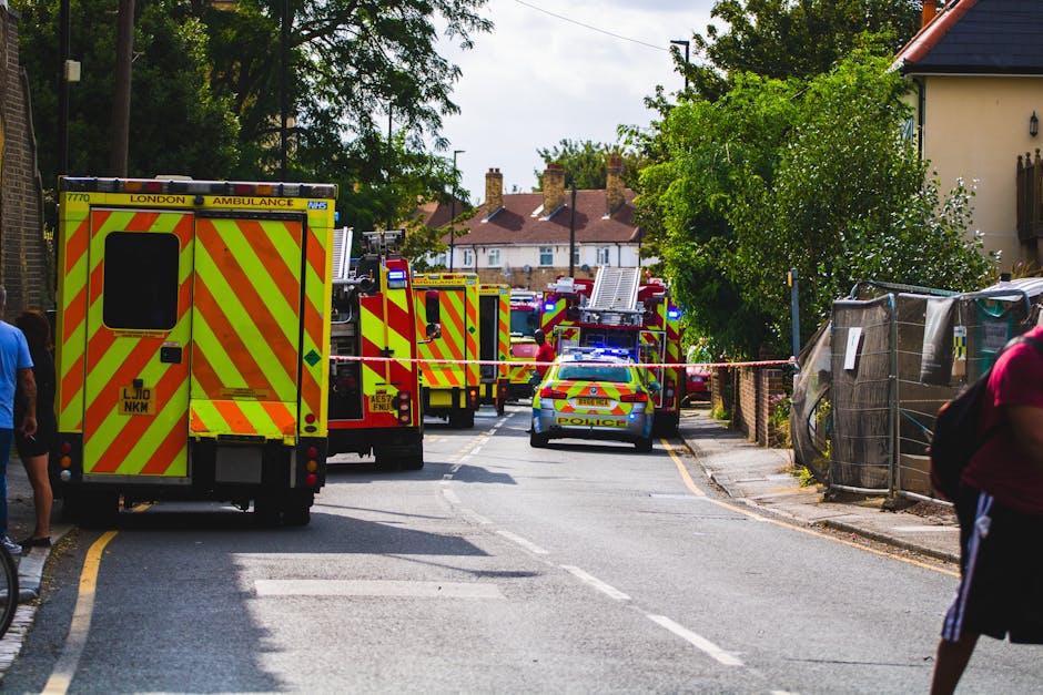 Emergency response scene on a residential street in Chadwell Heath with multiple fire trucks, a police car, and ambulance vehicles parked along the road. The fire trucks are yellow with red and orange diagonal striping, and their emergency lights are flashing. Several personnel and civilians are present, with some standing near the vehicles. The scene is cordoned off with red and white tape, blocking access to part of the street. Residential houses with chimneys and trees line both sides of the road, and the weather appears clear with daylight illuminating the area. The image captures a busy incident involving emergency responders, relevant to home relocations, safety, and logistics support, as managed by companies like Man with Van Chadwell Heath during urgent move situations.