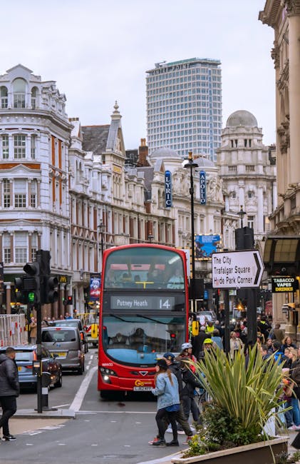 A busy urban street scene showing a red double-decker bus with route number 14 and destination Putney Heath, positioned in the center of the image as part of a home relocation and furniture transport process. The bus is surrounded by pedestrians on the pavement, some of whom are crossing the street, while others stand nearby. To the right, there are commercial shopfronts including a tobacconist, with black signage and yellow lettering, and pedestrians carrying cardboard boxes and plastic-wrapped items, indicating packing and moving activities. In the background, there are historic white-painted Victorian-style buildings with decorative architectural features, alongside a modern high-rise office tower, illustrating a typical city environment during moving or relocation tasks. The scene is outdoors during daytime, with clear lighting conditions, and the street is lined with lampposts, signage, and street furniture such as planters, all contributing to the context of urban furniture transport and house removals managed by Man with Van Chadwell Heath.