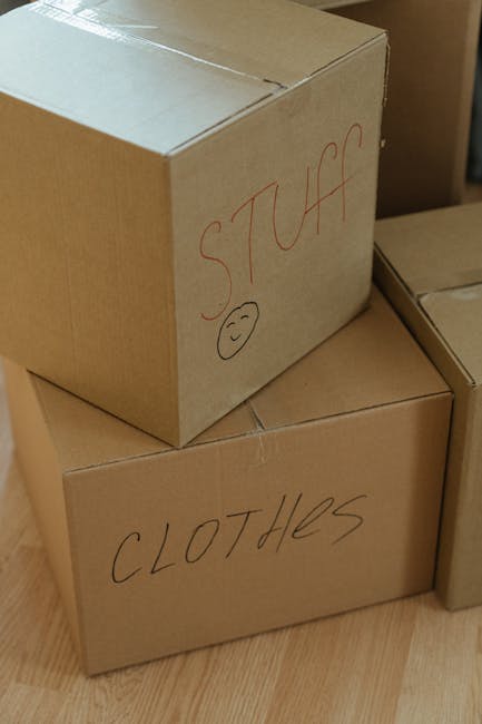 A group of three cardboard moving boxes is stacked on a wooden floor inside a property, with one box placed on top of two others. The top box is labeled 'STUFF' in red marker, accompanied by a small smiley face drawing. The two lower boxes are labeled 'CLOTHES' in black marker. The boxes are sealed with packing tape and show signs of careful packing for a home relocation. Nearby, part of another cardboard box is visible. The environment suggests an organized packing process, with the boxes prepared for transport by a moving service such as Man with Van Chadwell Heath, who specialise in packing and loading for house removals and furniture transport.