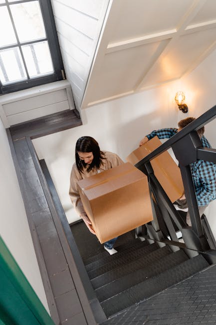 A woman with long dark hair, wearing a beige sweater, is carrying a medium-sized cardboard box up a staircase inside a residential property. She is walking on a black carpeted staircase with a black handrail and balustrade, with her head slightly bowed and focusing on the task. Behind her, a man wearing a blue checkered shirt is partially visible, also holding a cardboard box, on the same staircase. The staircase is adjacent to a white wall with a textured surface, and a window with a black frame is visible at the top, allowing natural light into the space. A small wall-mounted light fixture with a vintage-style bulb is also visible on the wall. The scene depicts a home relocation process involving packing and furniture transport, with the staircase being part of the interior environment where moving activities are taking place. The image supports content about house removals and delivery services provided by Man with Van Chadwell Heath.