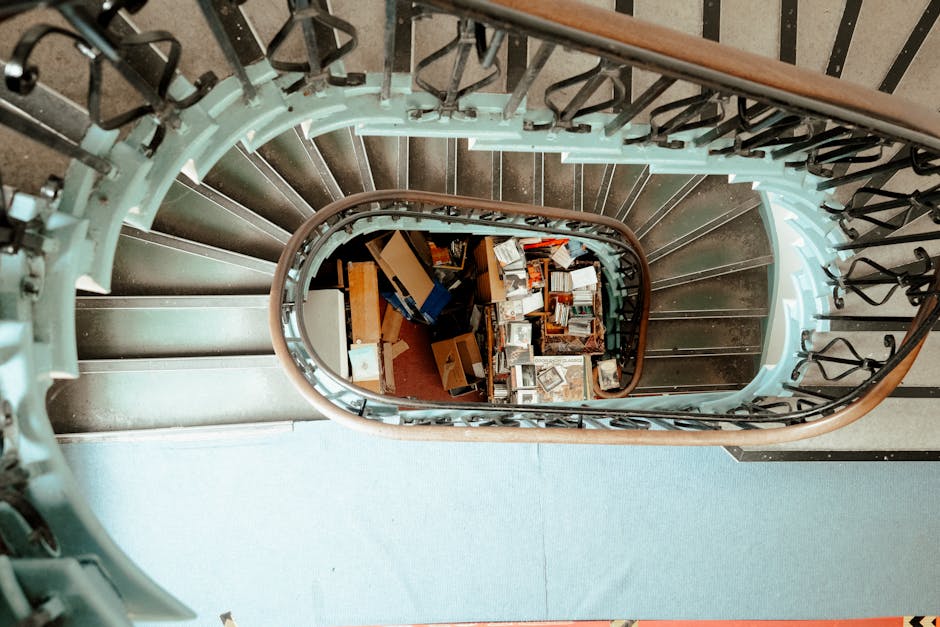 A view from the top of a spiral staircase showing the round wooden handrail and black metal balustrades. The staircase curves downward into a well-lit interior space with a light blue carpeted floor visible at the top. Below, on the landings, there are numerous cardboard boxes, some open, filled with books, packaging materials, and various household items, indicative of a house move or packing process. The boxes are arranged loosely, with some stacked and some scattered, with a few flat-packed furniture pieces or disassembled furniture parts among them. The environment appears to be an inside area of a home during a relocation, with the staircase leading to a lower floor. This image exemplifies furniture transport and packing activities related to house removals, as facilitated by Man with Van Chadwell Heath.
