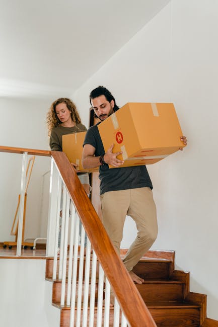 A woman with long dark hair, wearing a beige sweater, is carrying a medium-sized cardboard box up a staircase inside a residential property. She is walking on a black carpeted staircase with a black handrail and balustrade, with her head slightly bowed and focusing on the task. Behind her, a man wearing a blue checkered shirt is partially visible, also holding a cardboard box, on the same staircase. The staircase is adjacent to a white wall with a textured surface, and a window with a black frame is visible at the top, allowing natural light into the space. A small wall-mounted light fixture with a vintage-style bulb is also visible on the wall. The scene depicts a home relocation process involving packing and furniture transport, with the staircase being part of the interior environment where moving activities are taking place. The image supports content about house removals and delivery services provided by Man with Van Chadwell Heath.