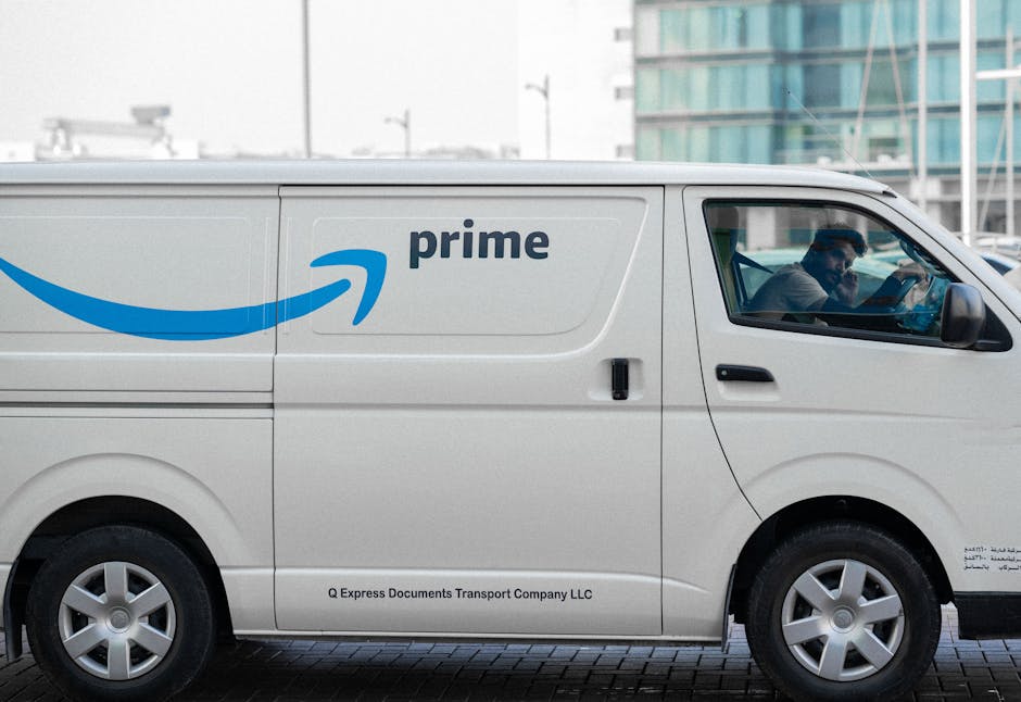 A white Amazon Prime delivery van parked outdoors on a paved surface with modern glass office buildings in the background. The van features the blue Amazon smile logo and the word 'prime' printed on t