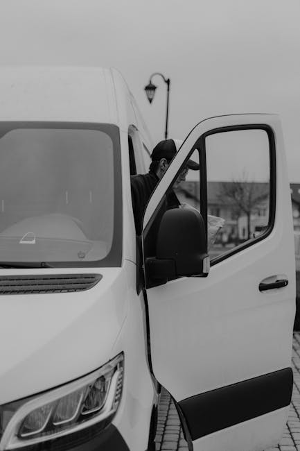 A white Amazon Prime delivery van parked outdoors on a paved surface with modern glass office buildings in the background. The van features the blue Amazon smile logo and the word 'prime' printed on t