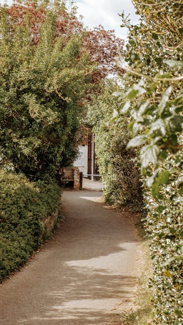 A narrow, paved pathway lined with dense, green foliage and trees on both sides, leading towards a residential street in Chadwell Heath. In the distance, there is a partial view of a brick property wi