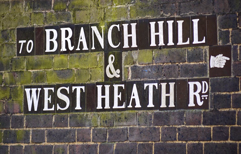 Close-up view of a brick wall with a street sign composed of individual rectangular tiles. The sign reads 'TO BRANCH HILL & WEST HEATH R.D.' with white lettering on a dark background, some tiles slightly weathered. To the right of the sign, there is a small white graphic of a pointing hand. The wall behind the sign appears aged, with moss and lichen growth giving it a slightly greenish tint. This street sign may be situated at an exterior or interior location relevant to a house relocation or moving services context, reflecting the environment where furniture and boxes might be loaded or unloaded.