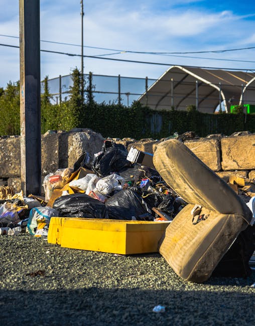 A pile of mixed household waste and rubbish bags, including black plastic garbage bags, cardboard boxes, and discarded furniture such as an old, worn-out car seat or armchair overturned on the ground. The waste is located outdoors on a paved surface beside a low stone wall, with a metal pole and power lines visible in the background. The scene is illuminated by natural daylight, suggesting daytime conditions. This image illustrates the type of bulky waste items that may be encountered during home relocation or furniture transport, which [COMPANY_NAME] in Chadwell Heath can assist with in their removals services. The setting reflects a typical outdoor waste collection area, with a focus on environmental and logistical considerations associated with moving and waste disposal.